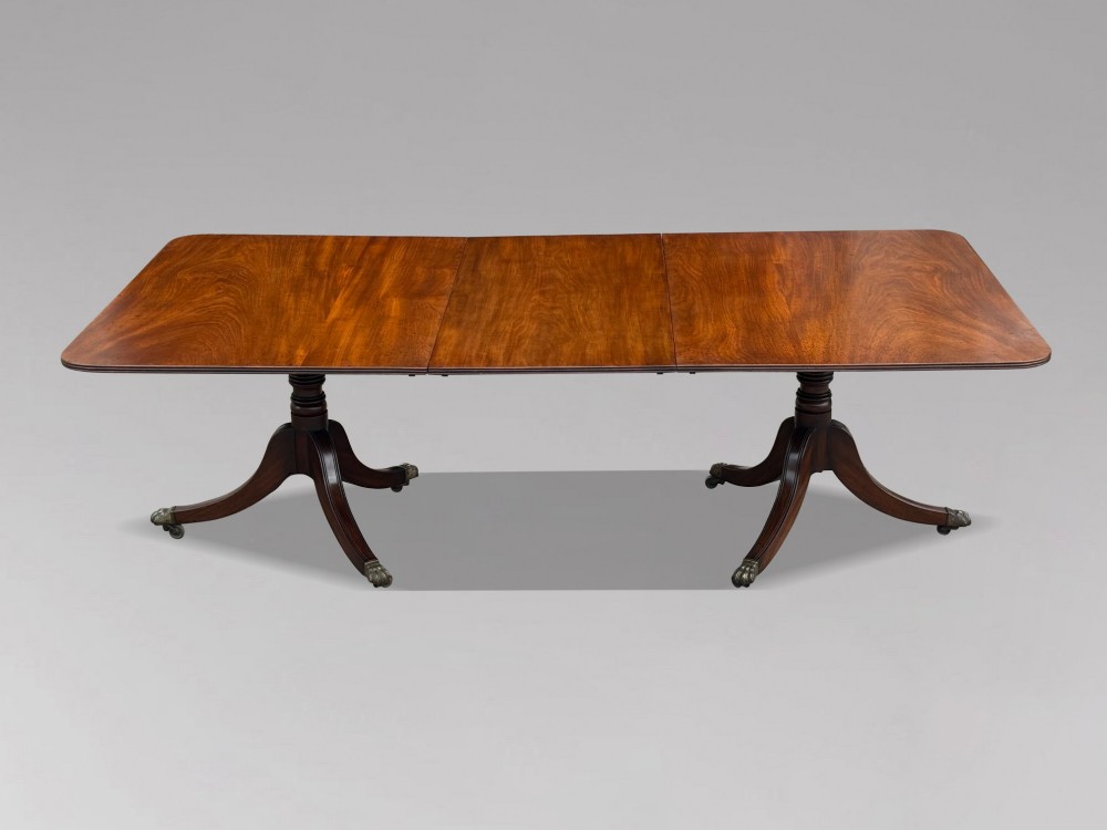 An early 19th-century Georgian mahogany twin pedestal dining table with a removable extension leaf, turned support pillars, and splayed legs ending in brass toe castors.