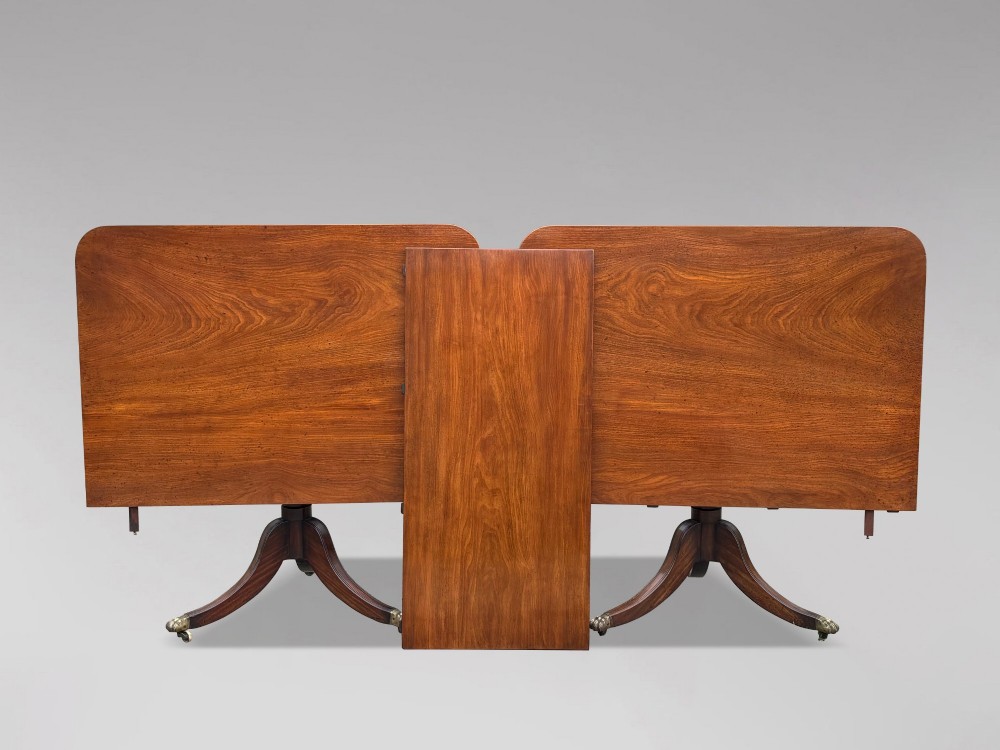 An early 19th-century Georgian mahogany twin pedestal dining table with a removable extension leaf, turned support pillars, and splayed legs ending in brass toe castors. View II