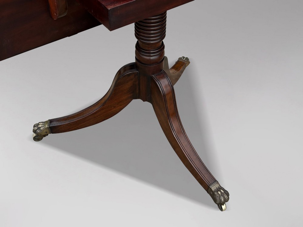 An early 19th-century Georgian mahogany twin pedestal dining table with a removable extension leaf, turned support pillars, and splayed legs ending in brass toe castors. View X