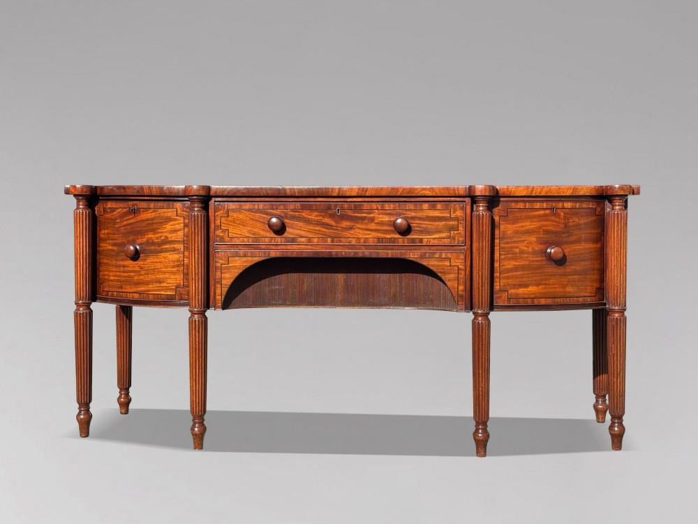 An antique 19th-century William IV mahogany sideboard featuring reeded fluted legs, line inlay, and a central drawer above an arched recess.
