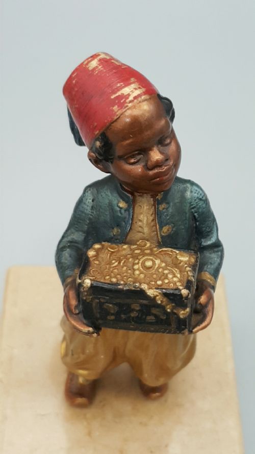 fig 1. An early 20th-century cold-painted bronze figure depicting a boy wearing a red fez and holding a treasure chest, mounted on a rectangular cream marble plinth.