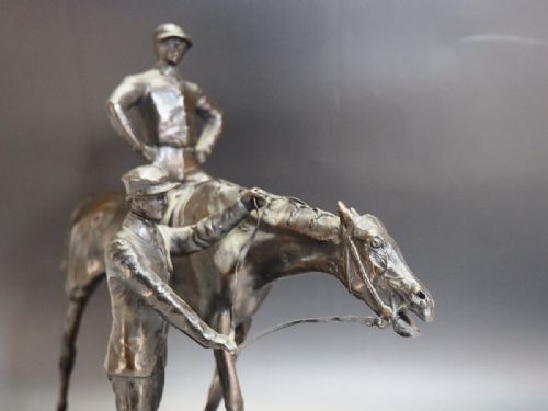 fig 1. A dark patinated bronze equestrian sculpture depicting a jockey mounted on a racehorse accompanied by a standing groom, set upon an oval green marble base.