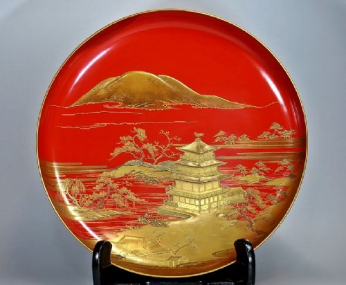 A large red lacquer Japanese sake cup decorated with gold maki-e motifs resting atop a dark brown wooden lacquer stand with a circular cutout. View IX