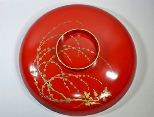 A large red lacquer Japanese sake cup decorated with gold maki-e motifs resting atop a dark brown wooden lacquer stand with a circular cutout. View XIV