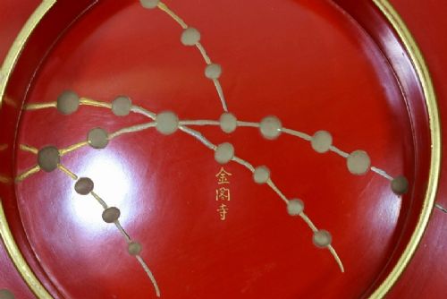 A large red lacquer Japanese sake cup decorated with gold maki-e motifs resting atop a dark brown wooden lacquer stand with a circular cutout. View XV