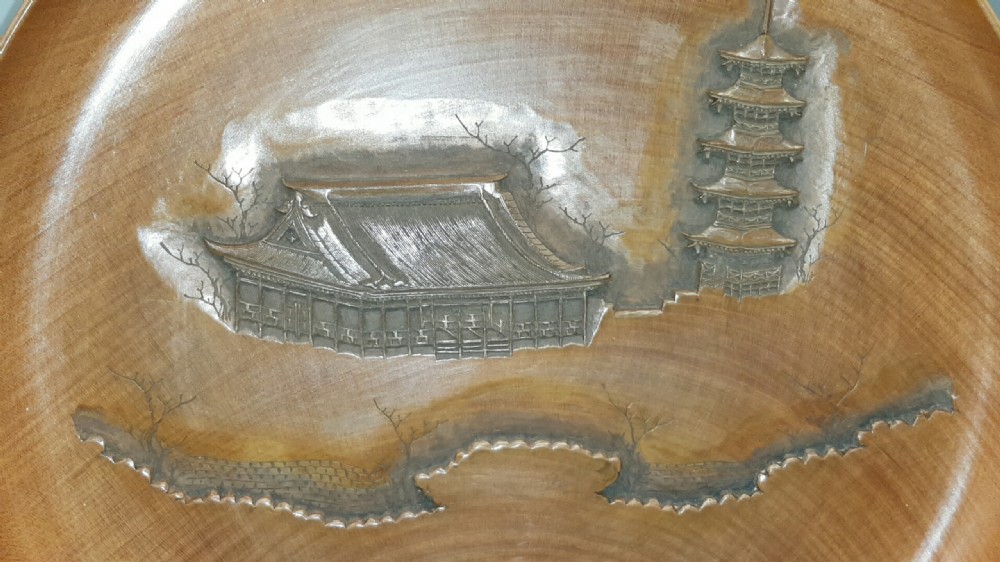 A stacked set of five shallow circular warm brown wood trays featuring low-relief carvings of traditional Japanese landscapes. View V