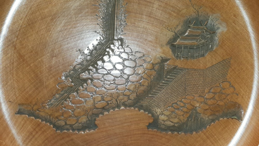 A stacked set of five shallow circular warm brown wood trays featuring low-relief carvings of traditional Japanese landscapes. View IX