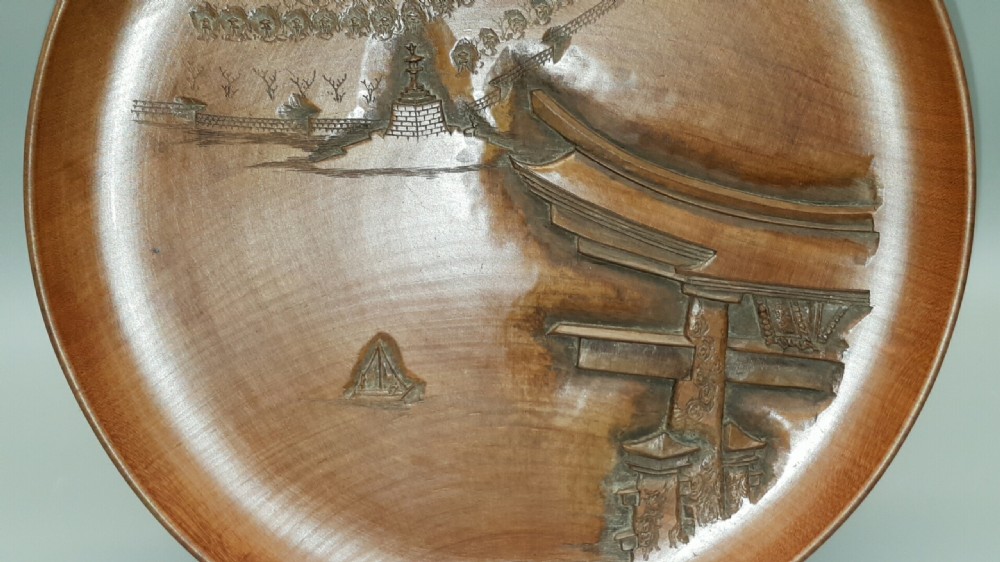 A stacked set of five shallow circular warm brown wood trays featuring low-relief carvings of traditional Japanese landscapes. View XI