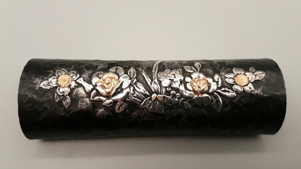 A cylindrical dark iron tea scoop featuring intricate silver and gold floral inlays across the surface.