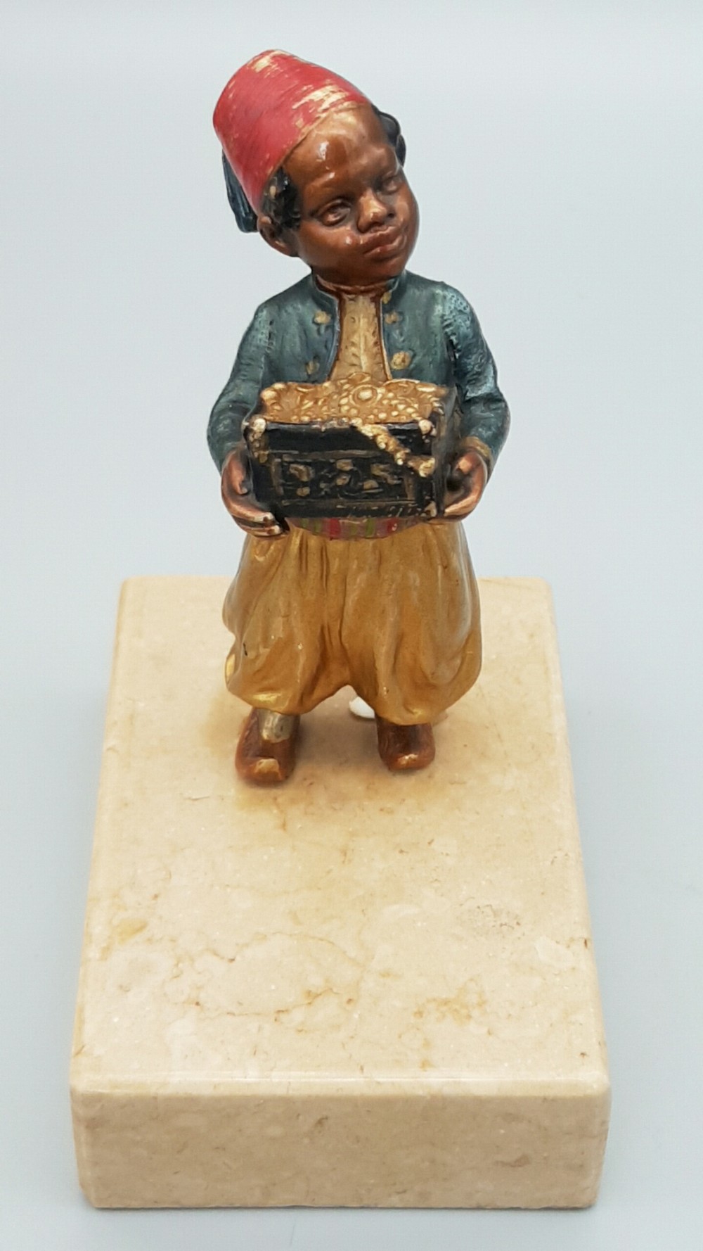 An early 20th-century cold-painted bronze figure depicting a boy wearing a red fez and holding a treasure chest, mounted on a rectangular cream marble plinth.