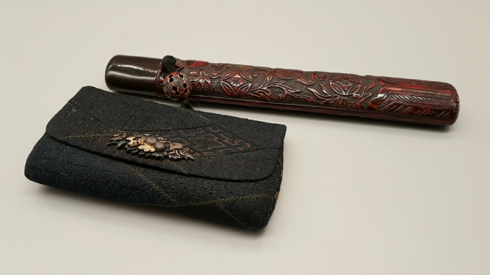 A Japanese Meiji period smoking ensemble featuring a dark woven silk tobacco pouch with a metal clasp beside a carved dark red-brown pipe case connected by a cord with an ojime bead.
