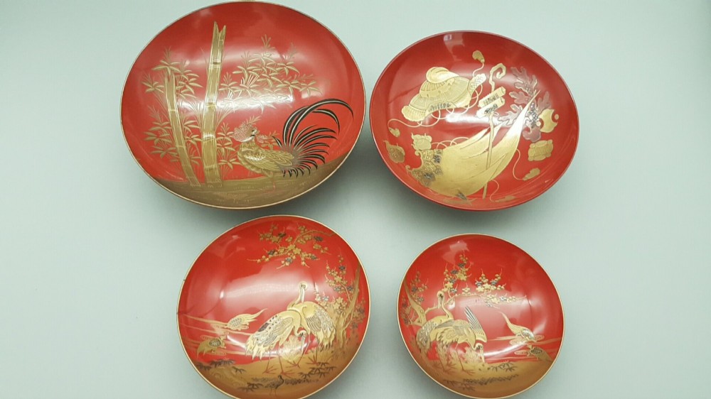 A set of four graduated Japanese red lacquer sake cups decorated with intricate gold maki-e illustrations including cranes, bamboo, and a rooster.