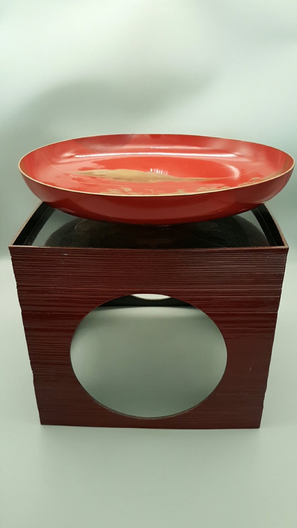 A large red lacquer Japanese sake cup decorated with gold maki-e motifs resting atop a dark brown wooden lacquer stand with a circular cutout.