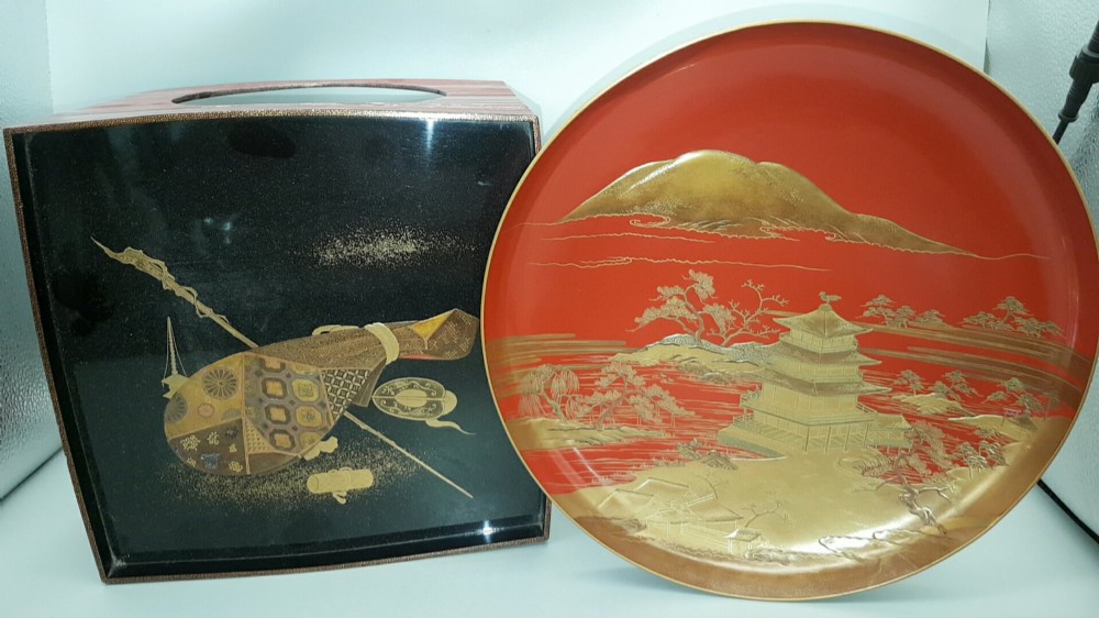 A large red lacquer Japanese sake cup decorated with gold maki-e motifs resting atop a dark brown wooden lacquer stand with a circular cutout. View II