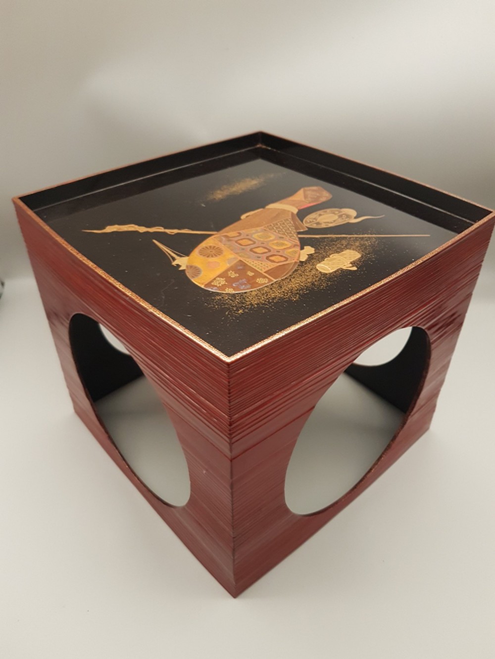 A large red lacquer Japanese sake cup decorated with gold maki-e motifs resting atop a dark brown wooden lacquer stand with a circular cutout. View III