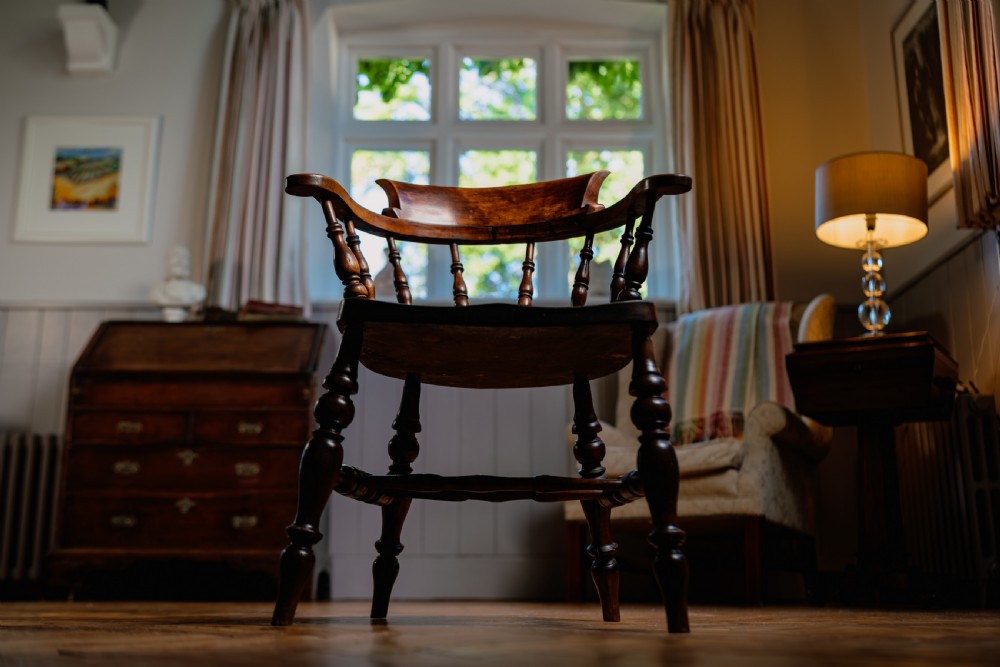 A dark brown wooden captains chair featuring a curved crest rail, turned spindles, a contoured saddle seat, and turned legs joined by stretchers. View II