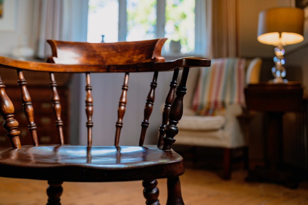 A dark brown wooden captains chair featuring a curved crest rail, turned spindles, a contoured saddle seat, and turned legs joined by stretchers. View III