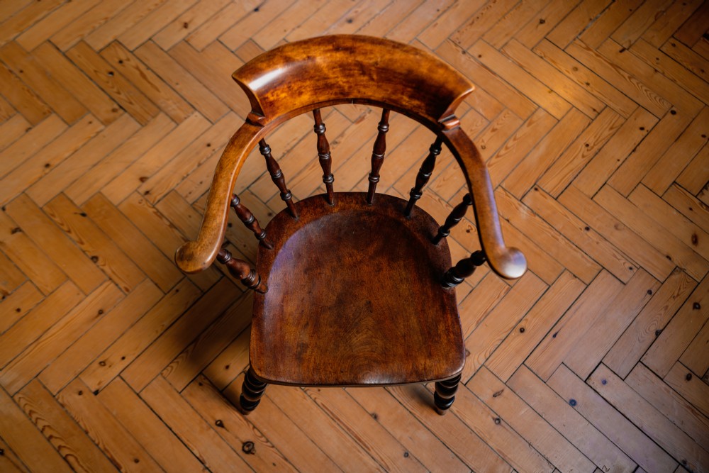A dark brown wooden captains chair featuring a curved crest rail, turned spindles, a contoured saddle seat, and turned legs joined by stretchers. View IV