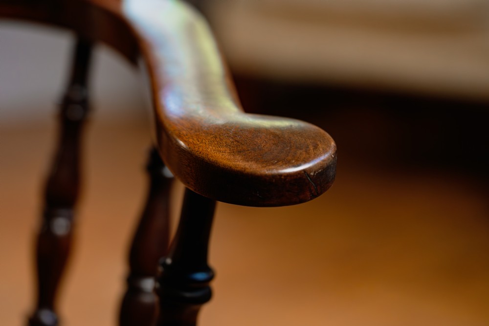 A dark brown wooden captains chair featuring a curved crest rail, turned spindles, a contoured saddle seat, and turned legs joined by stretchers. View V