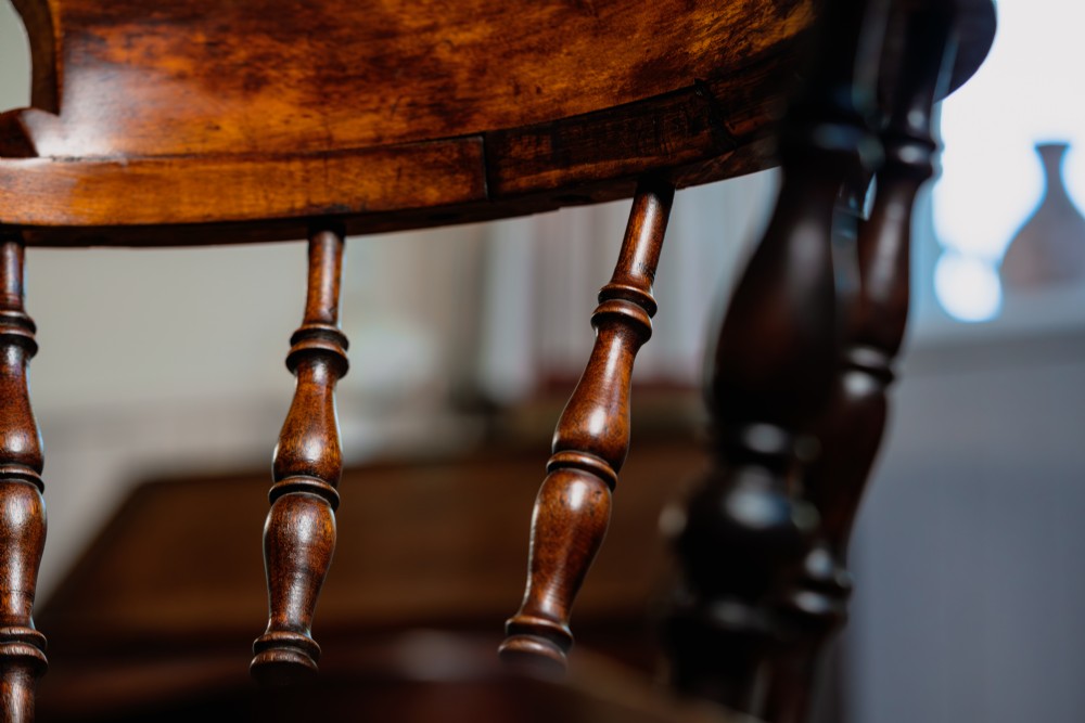 A dark brown wooden captains chair featuring a curved crest rail, turned spindles, a contoured saddle seat, and turned legs joined by stretchers. View VI