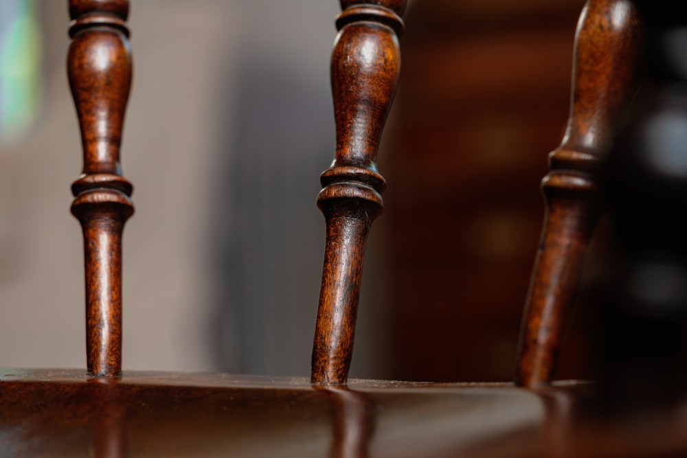 A dark brown wooden captains chair featuring a curved crest rail, turned spindles, a contoured saddle seat, and turned legs joined by stretchers. View VII