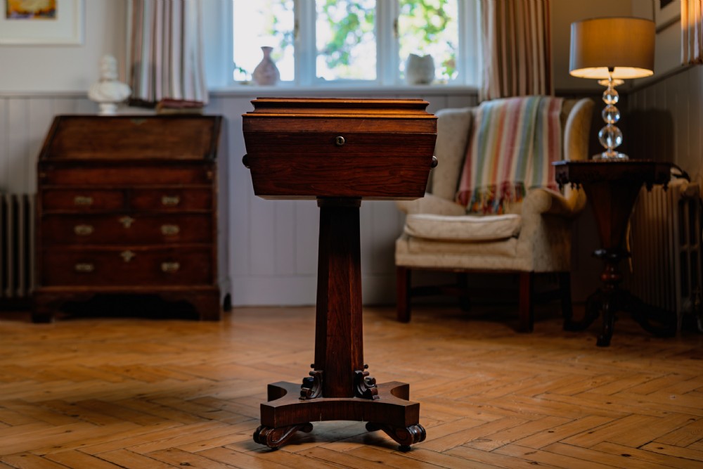 A Victorian rosewood teapoy featuring a rectangular hinged box top with a keyhole, supported by a tapered pedestal column on a four-pointed plinth base with scroll feet.