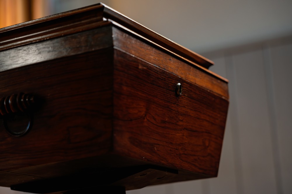A Victorian rosewood teapoy featuring a rectangular hinged box top with a keyhole, supported by a tapered pedestal column on a four-pointed plinth base with scroll feet. View VII