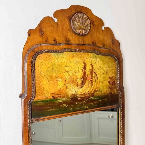 fig 1. A tall Queen Anne Revival yew wood wall mirror featuring a carved shell crest at the top, an upper panel painted with a sailing ship maritime scene, and a rectangular glass mirror below.