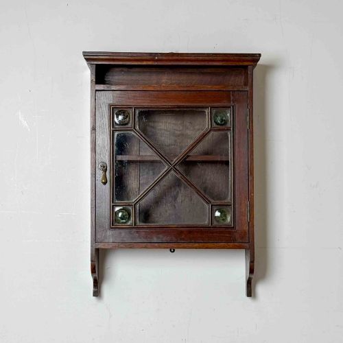 fig 1. A dark oak wall-mounted cabinet with a single glazed door featuring X-shaped glazing bars, brass drop handle, open top shelf, and shaped hanging brackets.