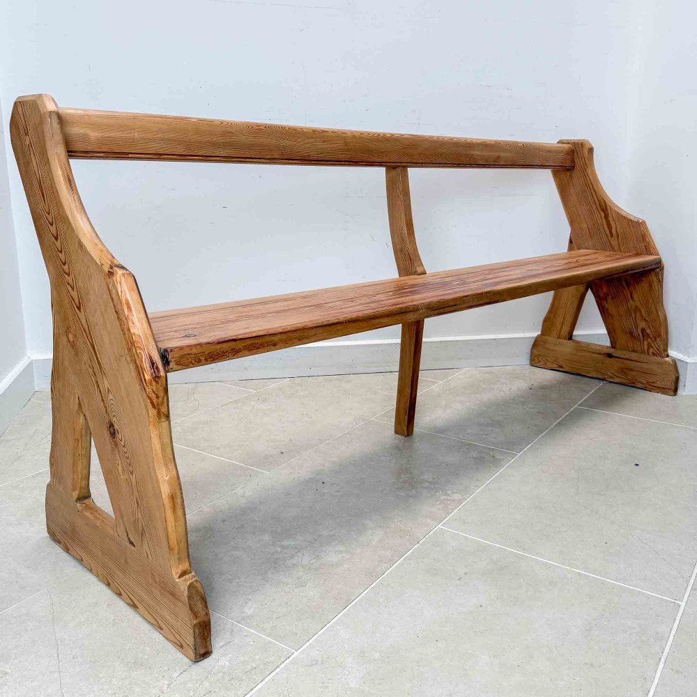 An antique pitch pine open back church pew featuring shaped side supports and a single central back support.