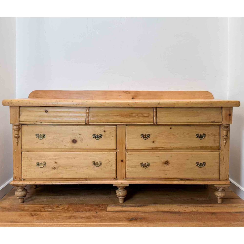 A Victorian natural pine dresser base featuring a low upstand back, seven drawers with metal drop handles, and turned bun feet.