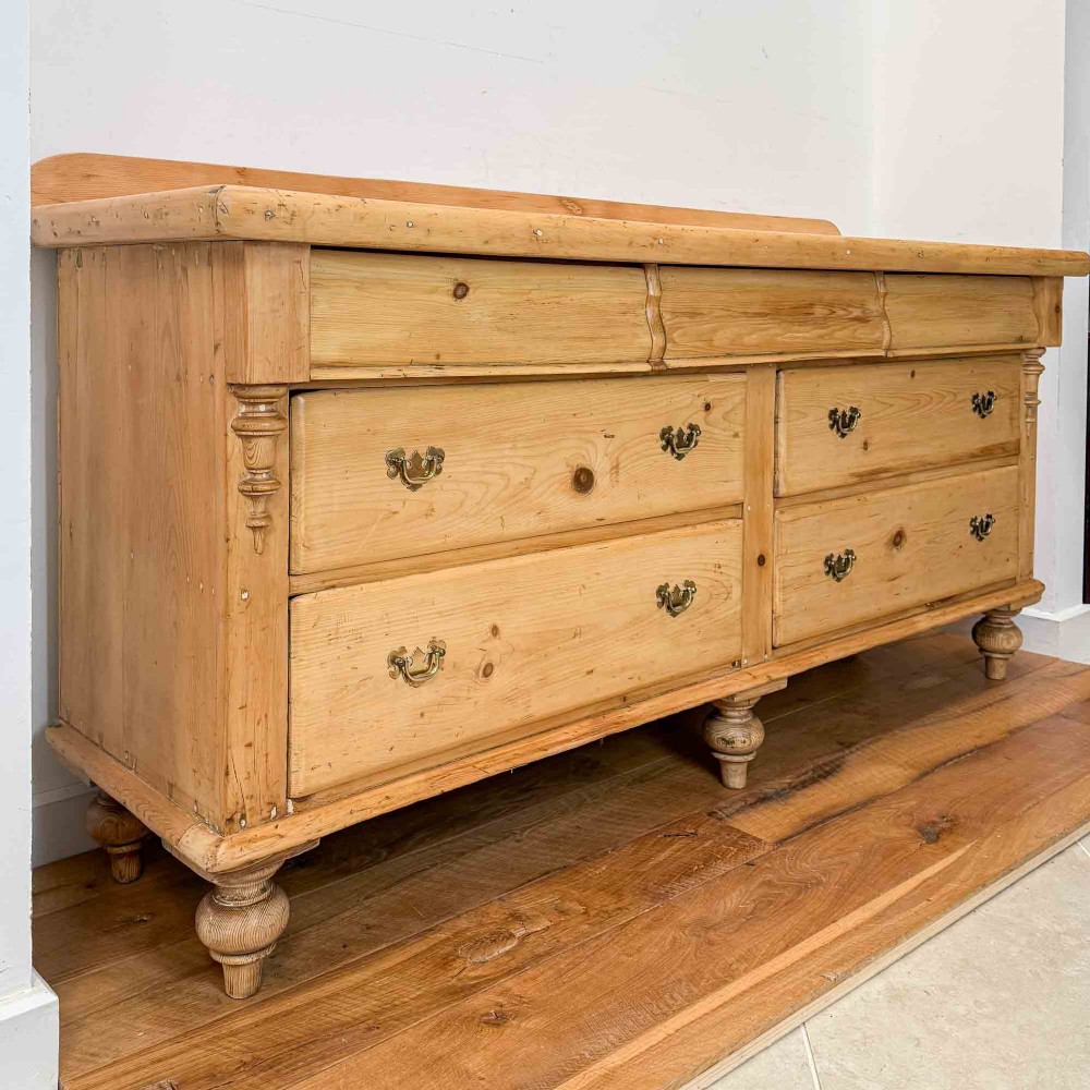 A Victorian natural pine dresser base featuring a low upstand back, seven drawers with metal drop handles, and turned bun feet. View II
