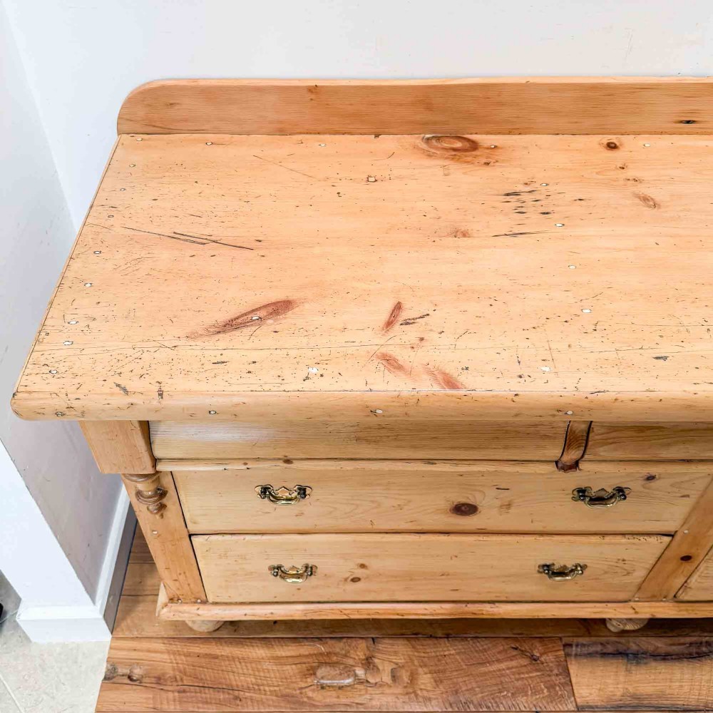 A Victorian natural pine dresser base featuring a low upstand back, seven drawers with metal drop handles, and turned bun feet. View XIII