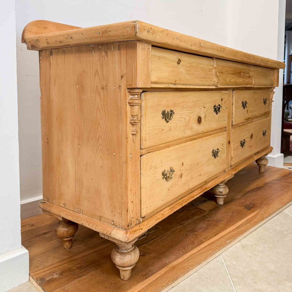 A Victorian natural pine dresser base featuring a low upstand back, seven drawers with metal drop handles, and turned bun feet. View XIV