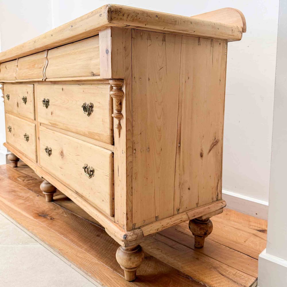 A Victorian natural pine dresser base featuring a low upstand back, seven drawers with metal drop handles, and turned bun feet. View XV