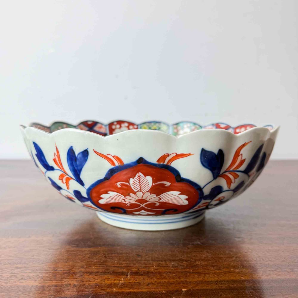 A shallow Japanese Imari porcelain bowl featuring a scalloped rim, hand-painted floral panels in cobalt blue, iron-red enamel, and green accents on a white porcelain ground. View VI