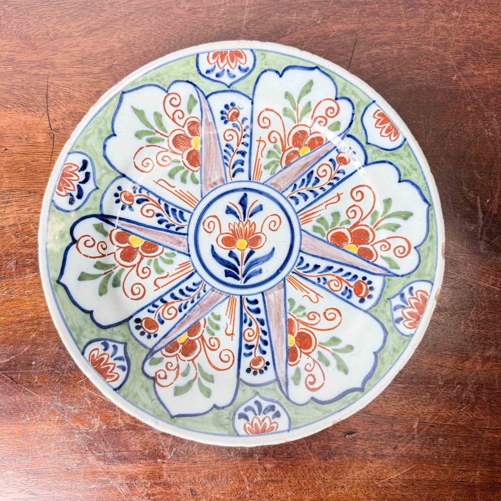 An antique circular tin-glazed earthenware plate decorated with hand-painted floral and geometric motifs in orange, cobalt blue, and brown over a pale green background.