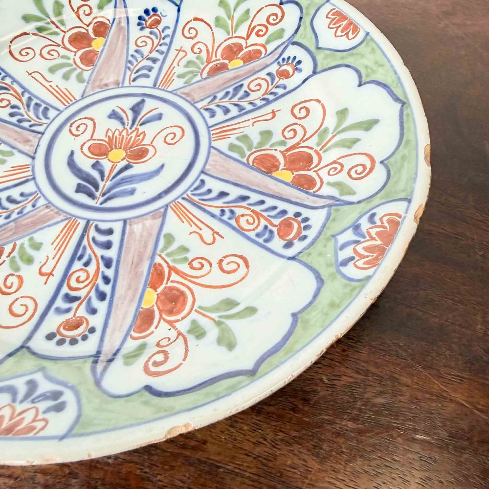 An antique circular tin-glazed earthenware plate decorated with hand-painted floral and geometric motifs in orange, cobalt blue, and brown over a pale green background. View II