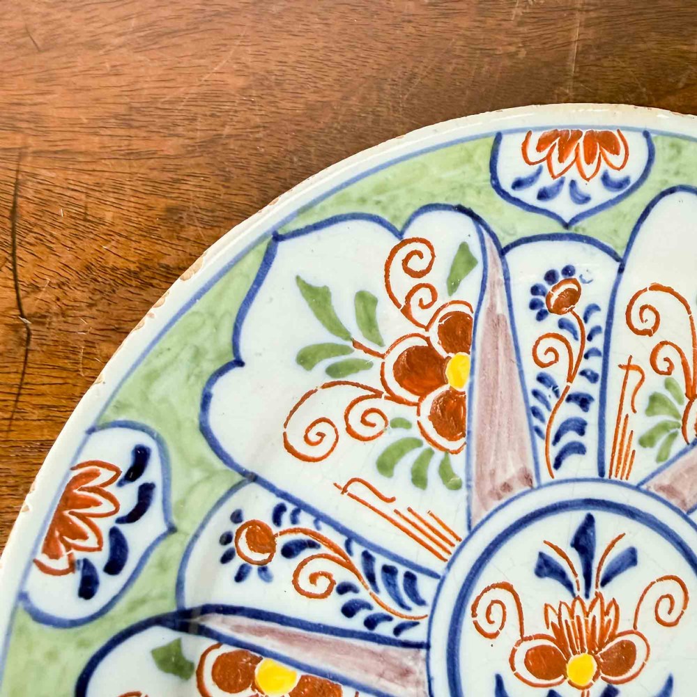 An antique circular tin-glazed earthenware plate decorated with hand-painted floral and geometric motifs in orange, cobalt blue, and brown over a pale green background. View III