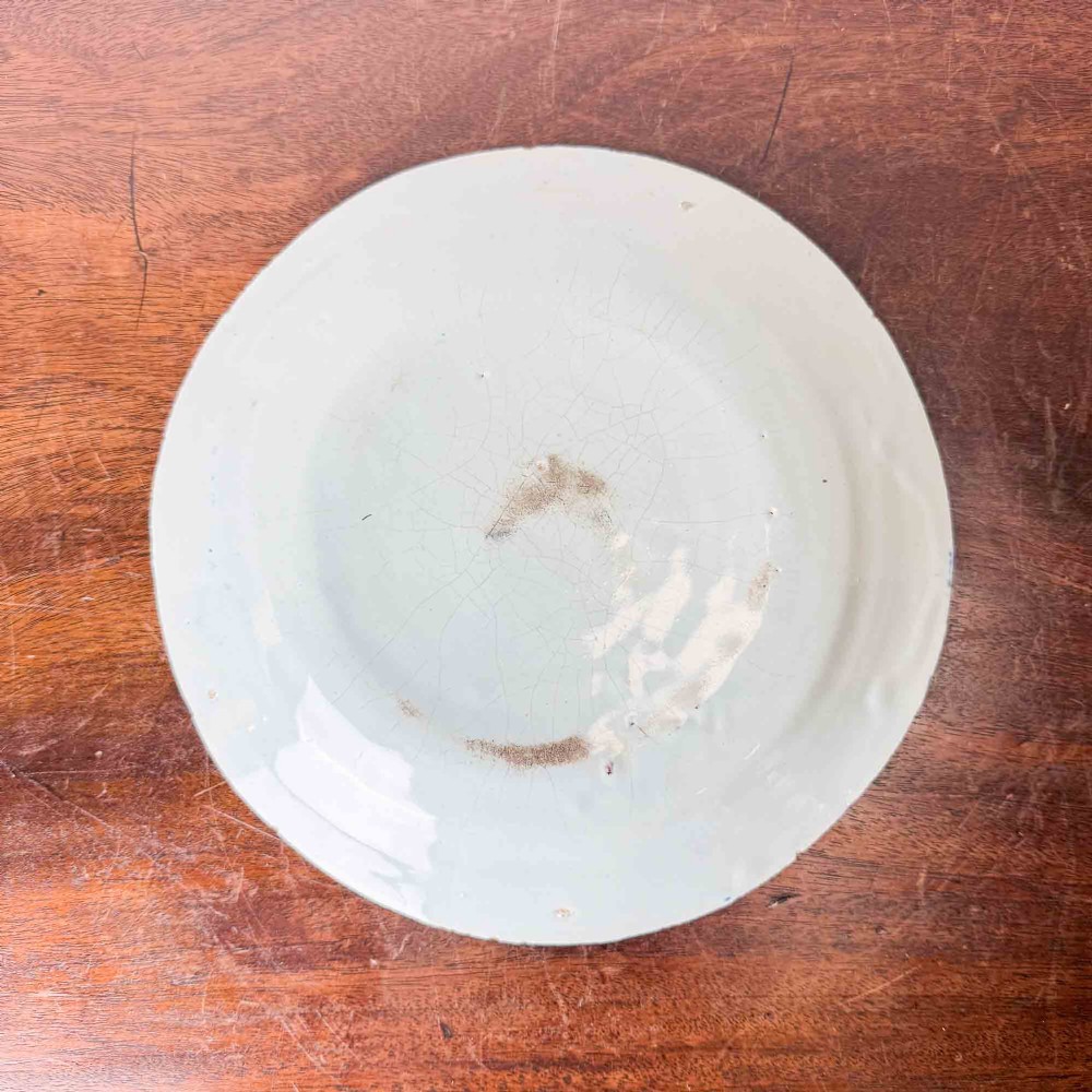 An antique circular tin-glazed earthenware plate decorated with hand-painted floral and geometric motifs in orange, cobalt blue, and brown over a pale green background. View IV