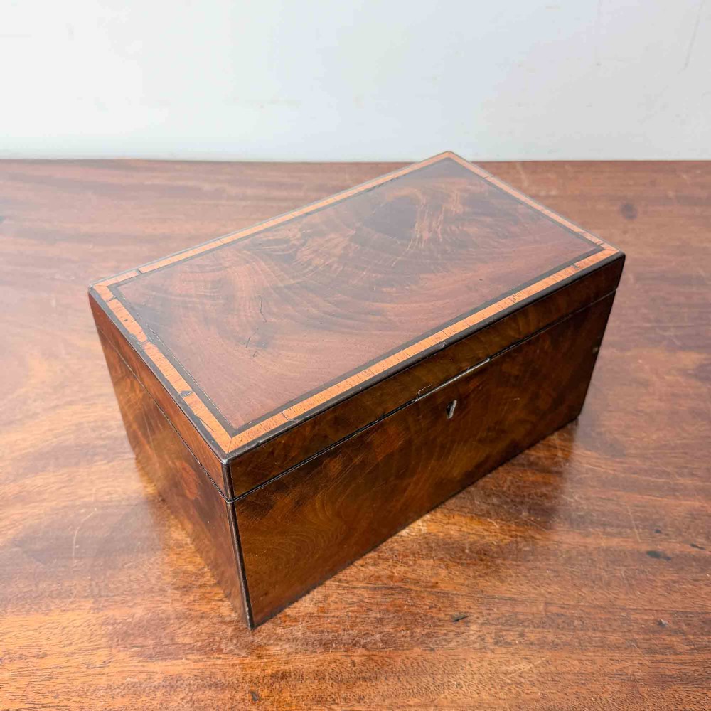 A rectangular early Victorian tea caddy crafted from flame mahogany veneer with crossbanded inlaid borders and a central metal keyhole escutcheon. View V