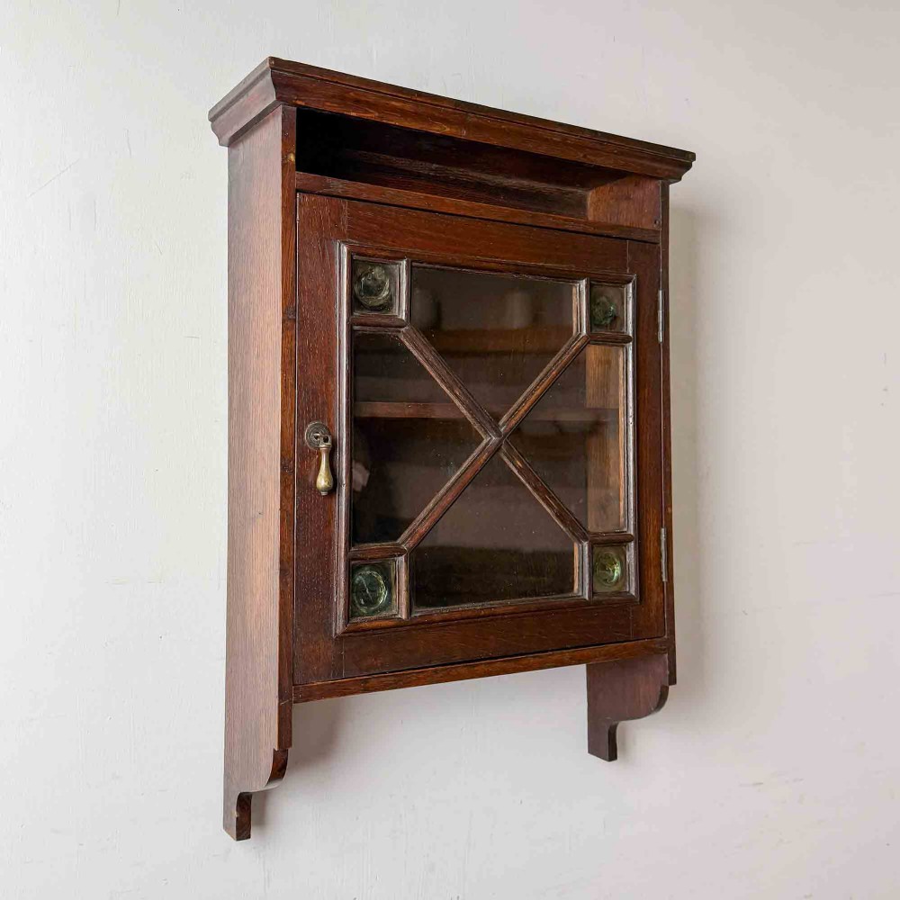 A dark oak wall-mounted cabinet with a single glazed door featuring X-shaped glazing bars, brass drop handle, open top shelf, and shaped hanging brackets.