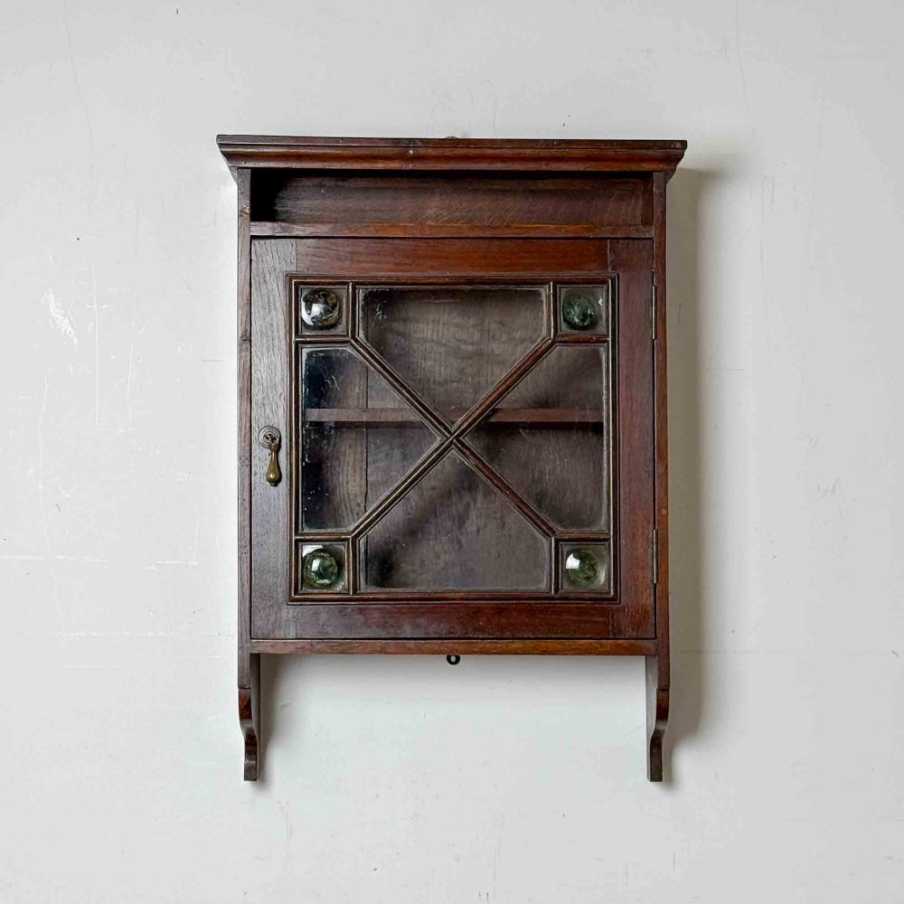 A dark oak wall-mounted cabinet with a single glazed door featuring X-shaped glazing bars, brass drop handle, open top shelf, and shaped hanging brackets. View II