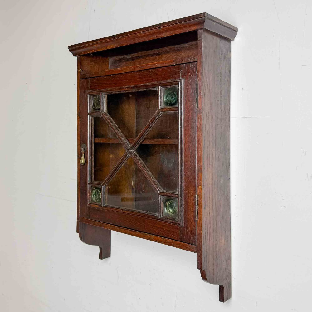 A dark oak wall-mounted cabinet with a single glazed door featuring X-shaped glazing bars, brass drop handle, open top shelf, and shaped hanging brackets. View III