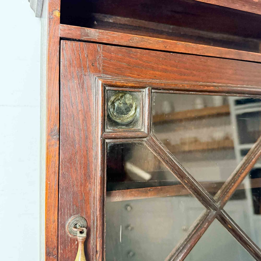 A dark oak wall-mounted cabinet with a single glazed door featuring X-shaped glazing bars, brass drop handle, open top shelf, and shaped hanging brackets. View IV