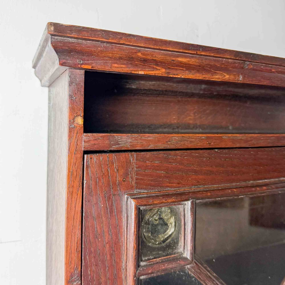 A dark oak wall-mounted cabinet with a single glazed door featuring X-shaped glazing bars, brass drop handle, open top shelf, and shaped hanging brackets. View VI