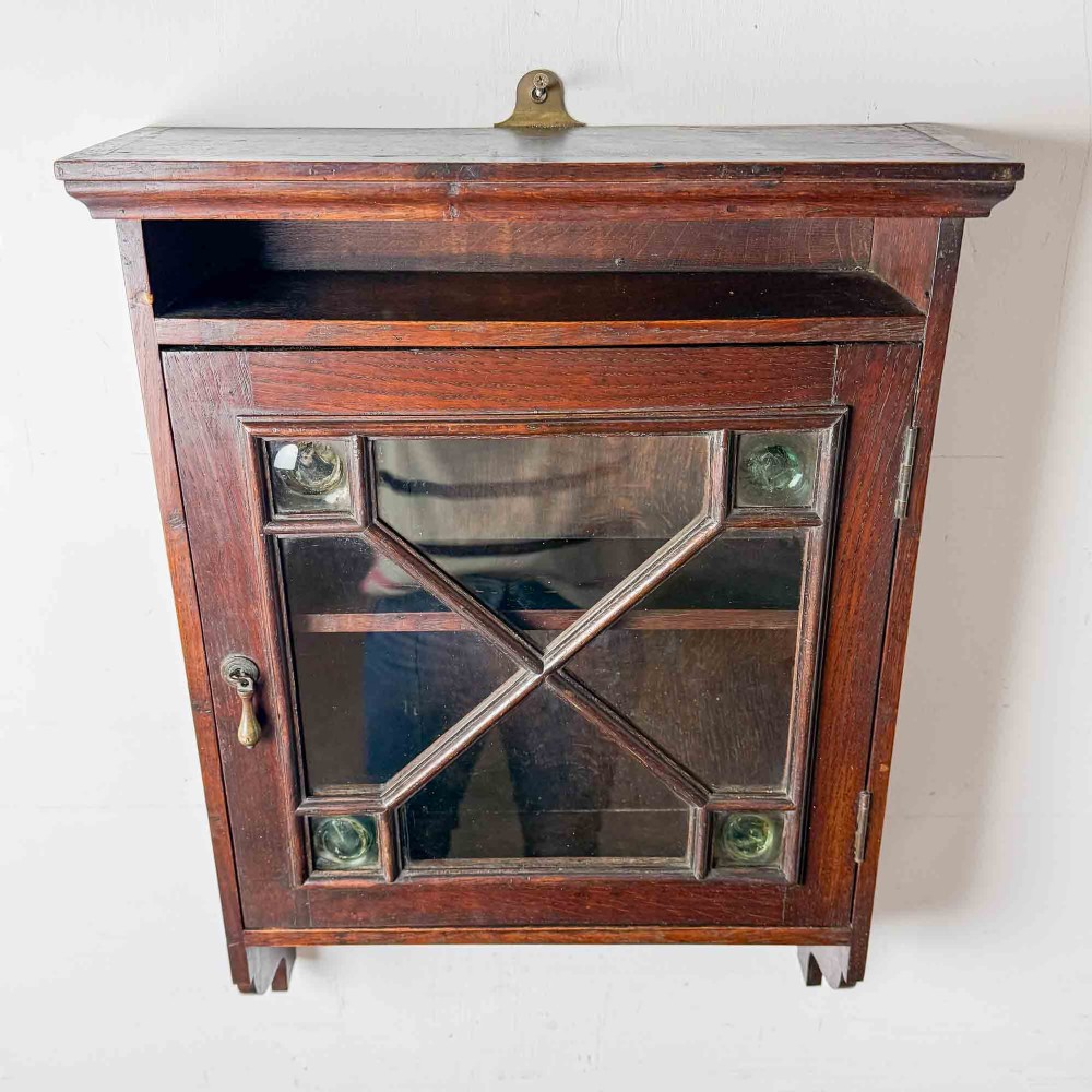 A dark oak wall-mounted cabinet with a single glazed door featuring X-shaped glazing bars, brass drop handle, open top shelf, and shaped hanging brackets. View VII
