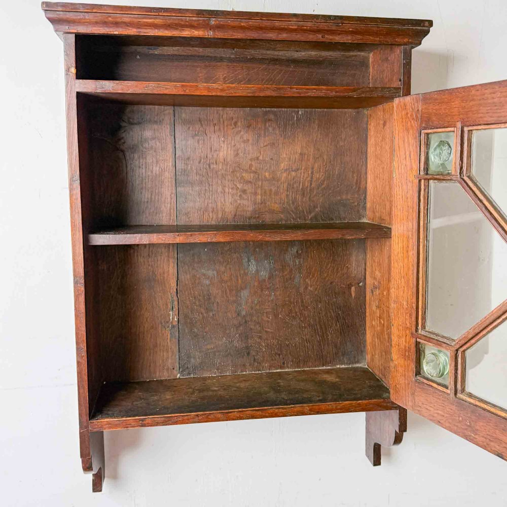 A dark oak wall-mounted cabinet with a single glazed door featuring X-shaped glazing bars, brass drop handle, open top shelf, and shaped hanging brackets. View VIII