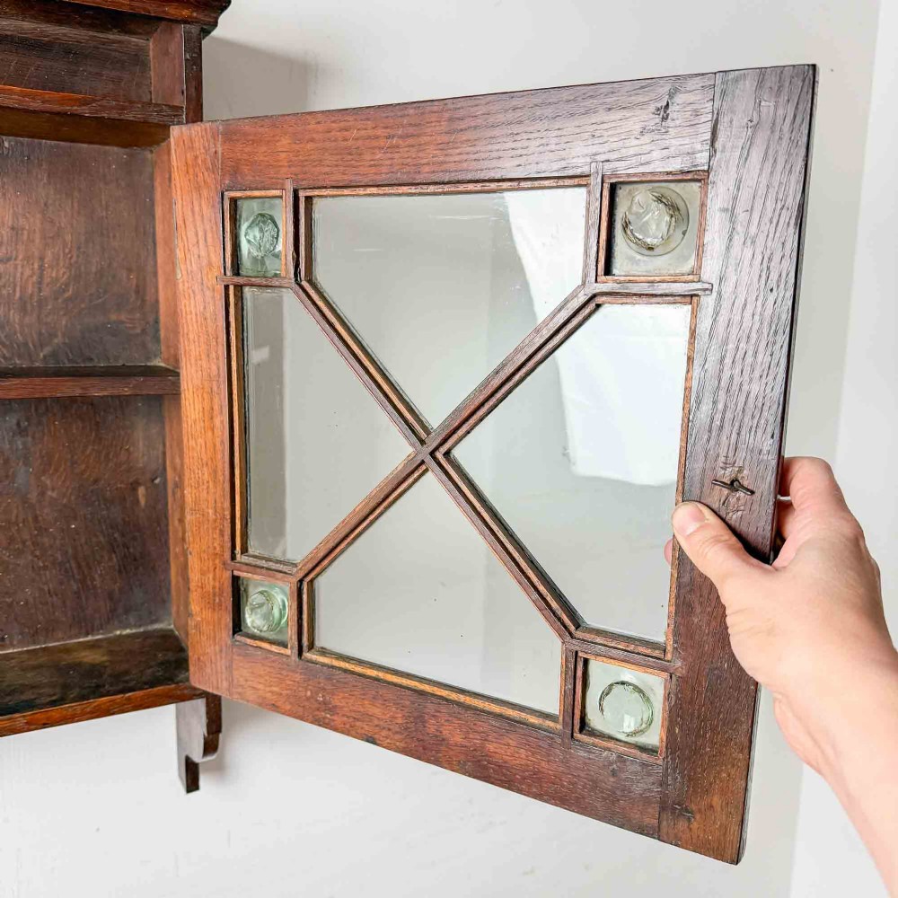 A dark oak wall-mounted cabinet with a single glazed door featuring X-shaped glazing bars, brass drop handle, open top shelf, and shaped hanging brackets. View IX