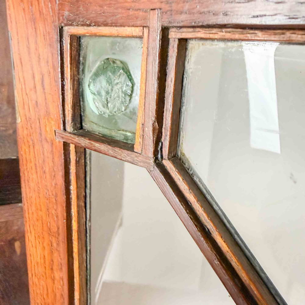A dark oak wall-mounted cabinet with a single glazed door featuring X-shaped glazing bars, brass drop handle, open top shelf, and shaped hanging brackets. View X